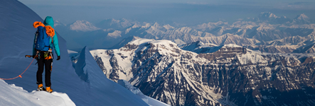 A climber wearing a blue jacket and carrying a backpack with coiled orange rope stands on a steep snow slope, looking away from the camera toward a dramatic horizon of endless mountain ranges under a blue sky.