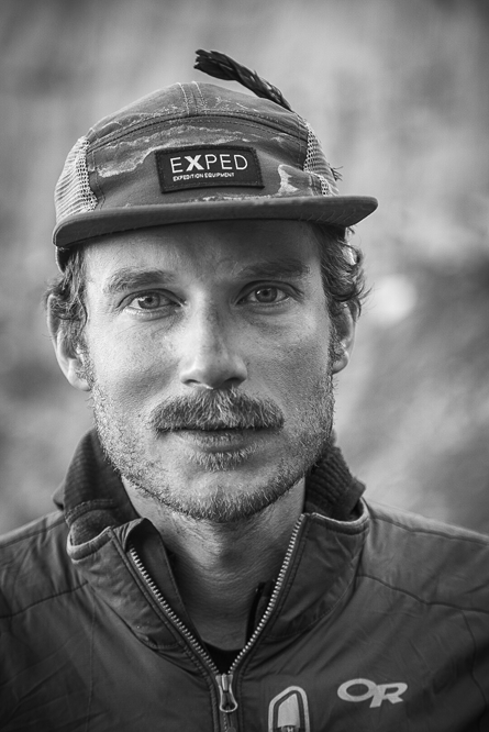 A black and white portrait of alpinist Graham Zimmerman looking directly at the camera. He features a mustache and stubble, and is wearing a worn cap with an EXPED logo patch and a zipped jacket with an Outdoor Research logo on the chest.